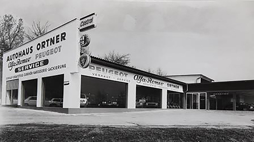 Schwarz-Weiß-Aufnahme: das Autohaus Ortner in der Piccostraße mit den Schriftzügen Alfa Romeo, Peugeot und Service, Fahrzeuge hinter der Glasfront