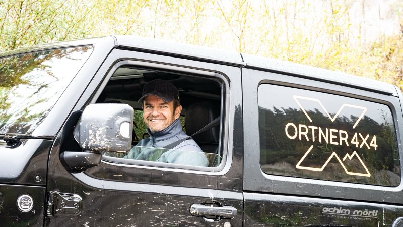 Ein Fahrer am Steuer eines schwarzen Jeep Wrangler mit Ortner-4x4-Scheibenbeschriftung, das Fahrzeug ist vom Gelände verschmutzt
