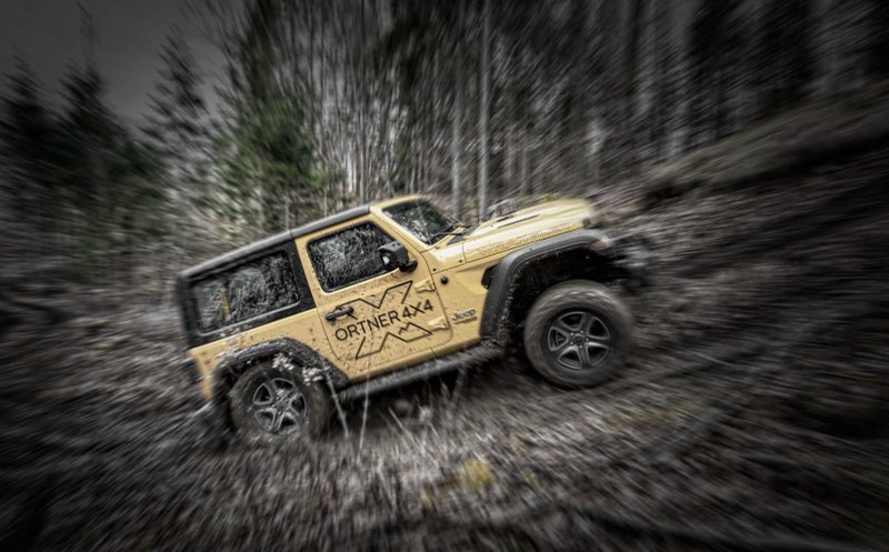 Ein sandfarbener Jeep Wrangler mit Ortner-4x4-Beschriftung fährt durch tiefen Schlamm im Wald, das Bild ist von der Bewegung verwischt